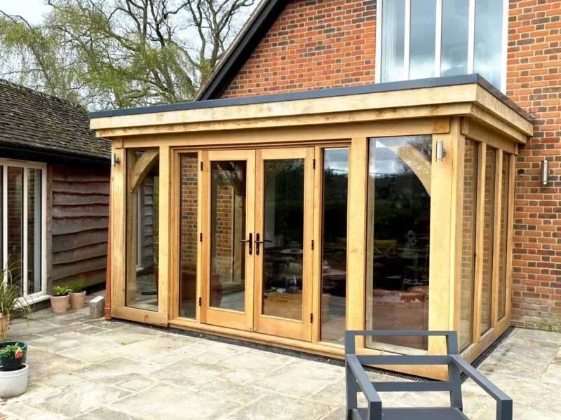 A modern oak frame orangery with a flat roof and large glass doors, attached to a brick house, opening onto a paved patio area.