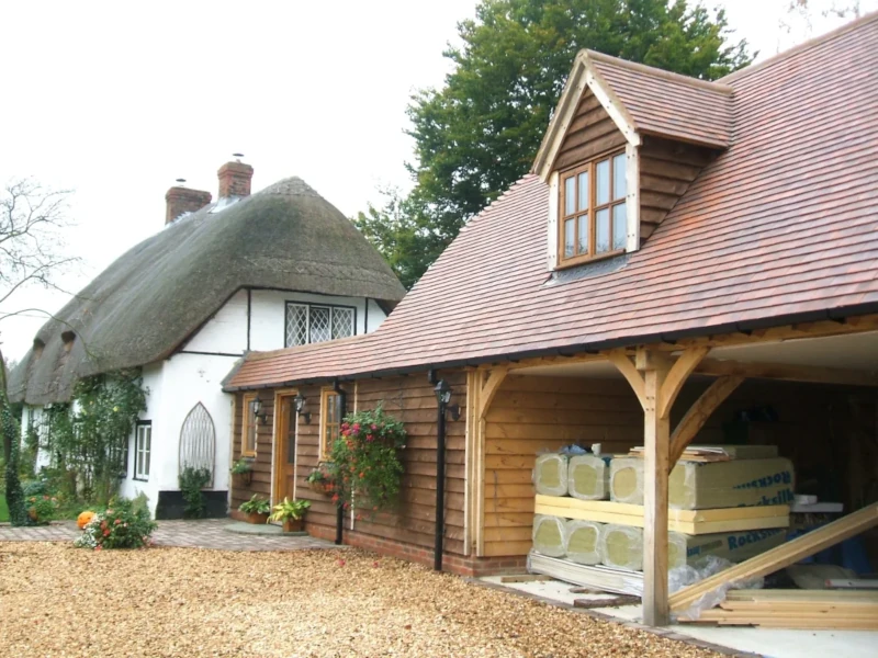 A newly built oak frame extension with exposed beams and a carport stands alongside a picturesque thatched cottage.