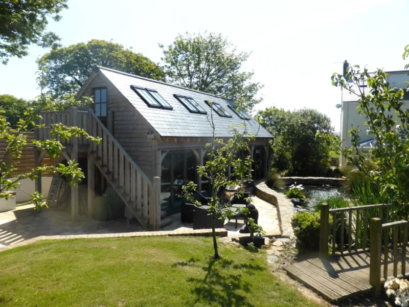 A picturesque oak framed pool house garden studio with a slate roof, multiple skylights, and an exterior staircase leading to a balcony. The studio overlooks a landscaped garden with a pond and a small bridge.