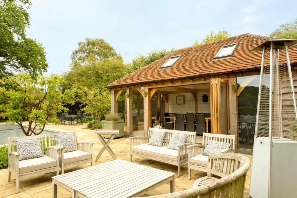 A luxurious oak framed pool house with open bi-fold doors revealing a spacious interior and a view of a swimming pool.