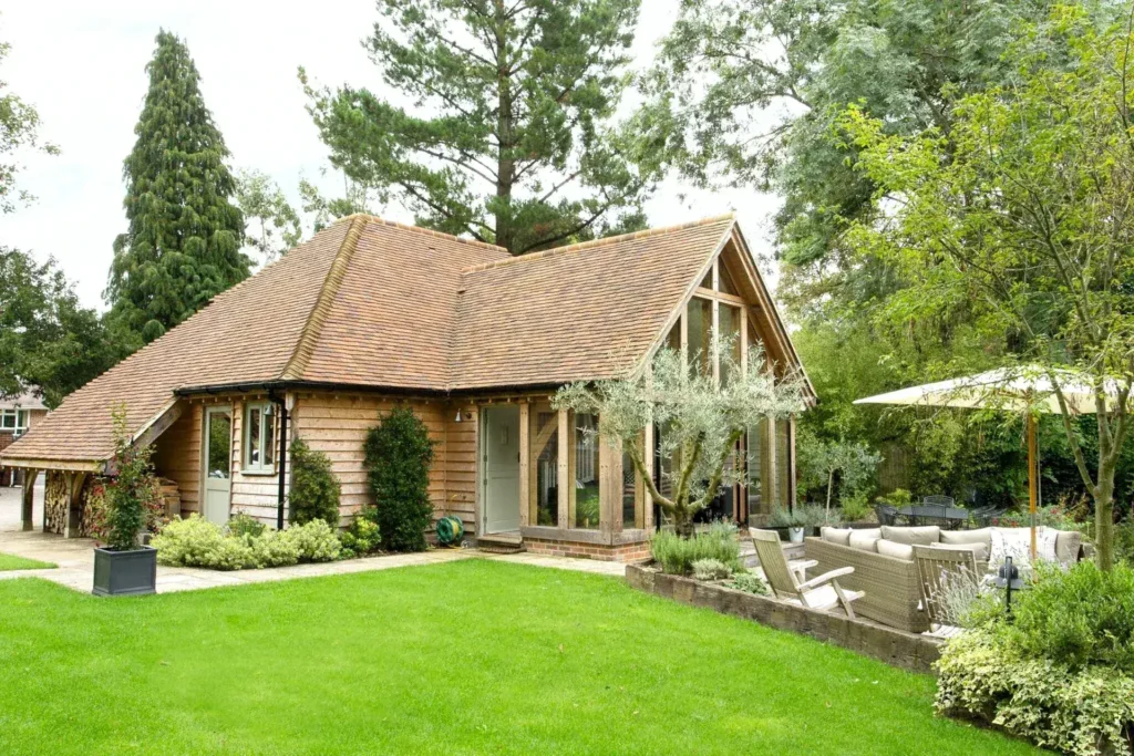 A charming oak framed annexe nestled in a garden setting, featuring a glazed gable and a patio area with outdoor seating.