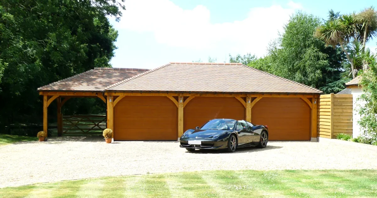 Bespoke Oak Framed Garages Dorset Luxury bespoke oak framed garages Dorset: Four bay garage with tiled roof and wooden doors, featuring a black Ferrari parked outside on gravel driveway