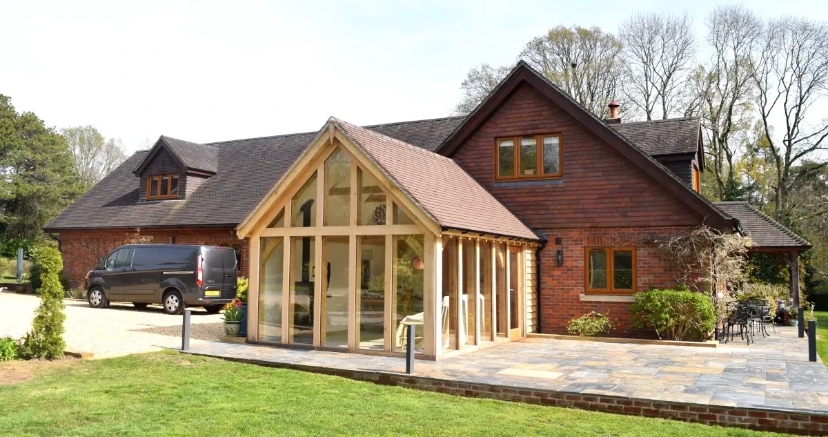 Our bespoke oak framed sunrooms can be designed as stunning extensions with floor-to-ceiling glazing. This oak garden rooms is attached to a traditional brick and timber country home in the UK, featuring natural oak beams and stone patio