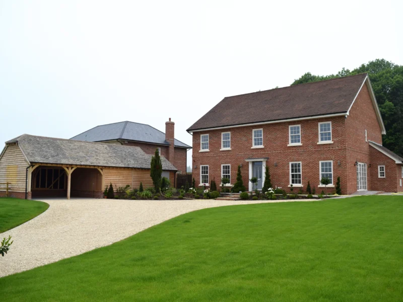 Search our bespoke oak buildings: an oak frame garage & carport alongside a red brick georgian-style house with landscaped gardens and gravel driveway