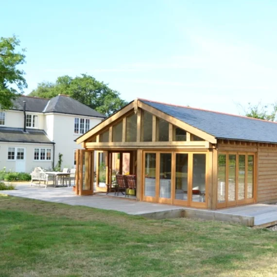 Oak frame garden annexe with full-height glazing and cedar cladding alongside traditional white house in rural uk garden setting