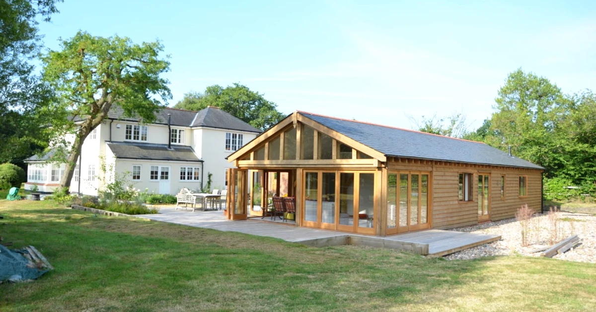 Oak frame garden annexe with full-height glazing and cedar cladding alongside traditional white house in rural uk garden setting