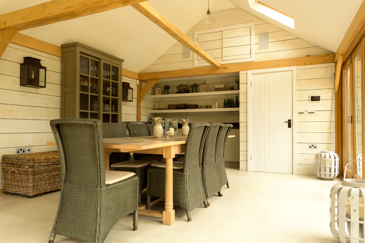 Vaulted dining room in oak frame annexe featuring painted tongue and groove walls, exposed beams, built-in storage and sage green rattan dining chairs