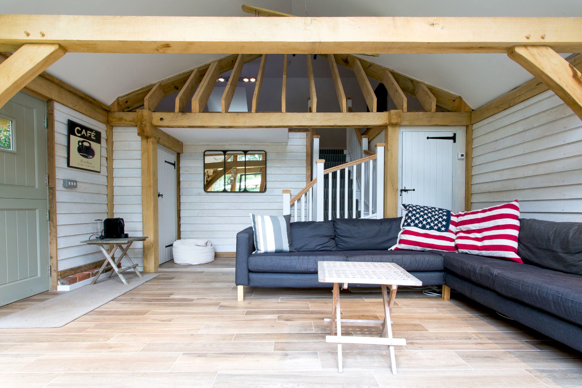 Modern oak frame annexe living room featuring vaulted ceiling, exposed beams, painted cladding, grey corner sofa and wood-effect flooring with mezzanine stairs