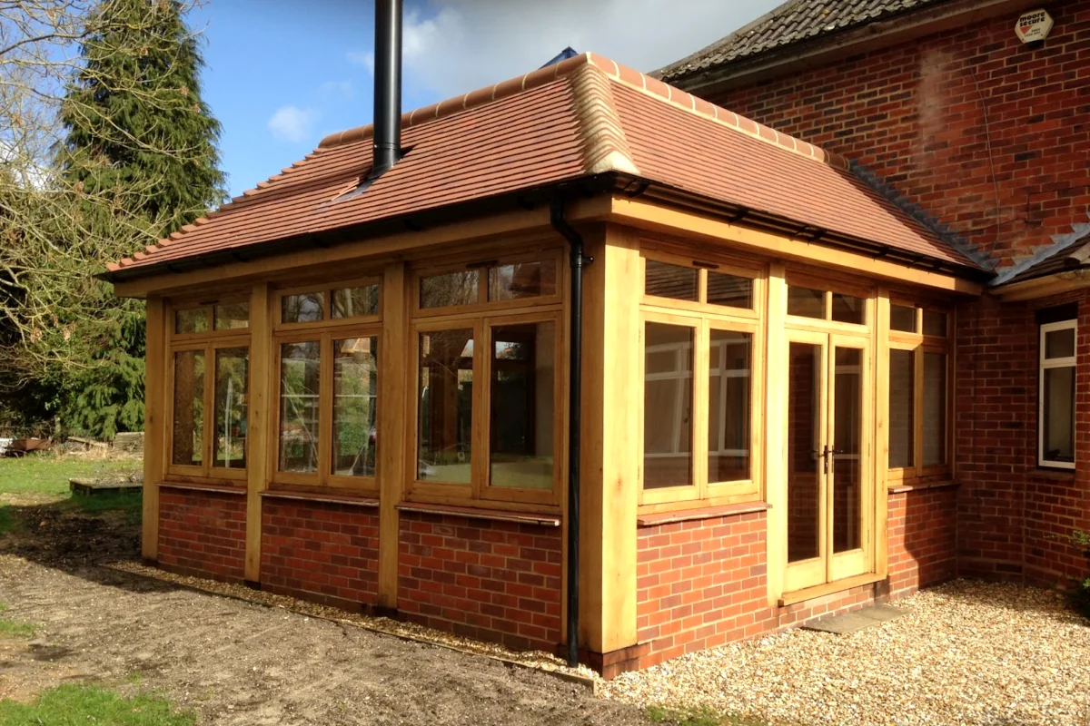 Oak frame construction project showing garden room extension with clay tile roof, full-height glazing, brick plinth and oak frame windows, seamlessly connected to red brick house