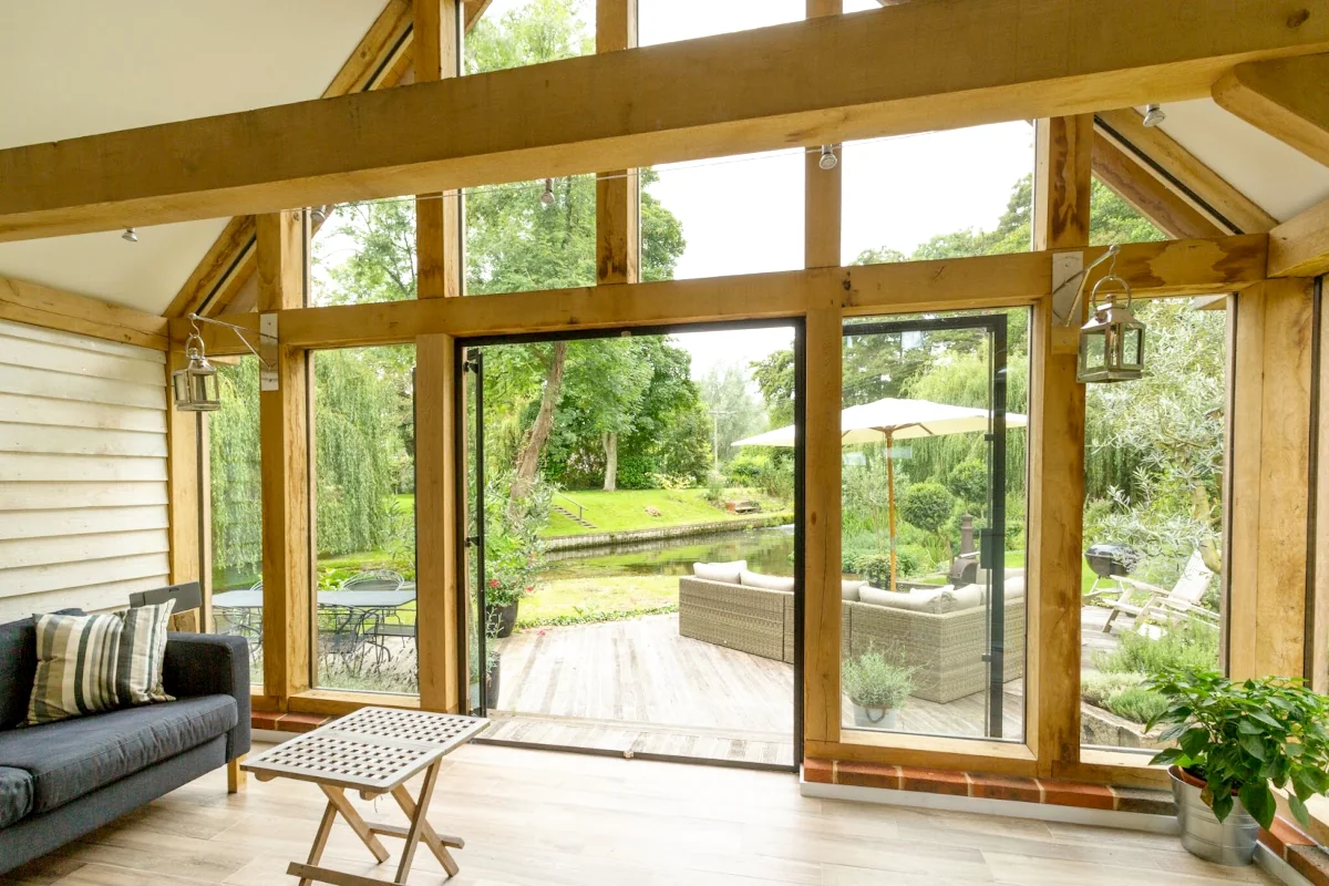 Interior view of an oak frame garden room extension with exposed beams, full-height glazing, bifold doors opening onto garden decking with pond view, showing natural integration with existing home