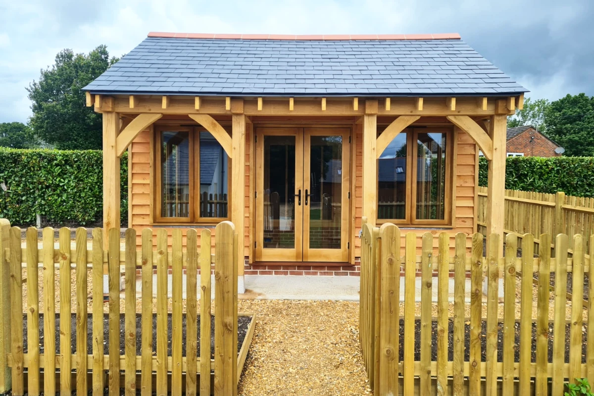 Traditional oak frame garden office with slate roof, french doors, and picket fence, featuring handcrafted timber posts and natural cedar cladding