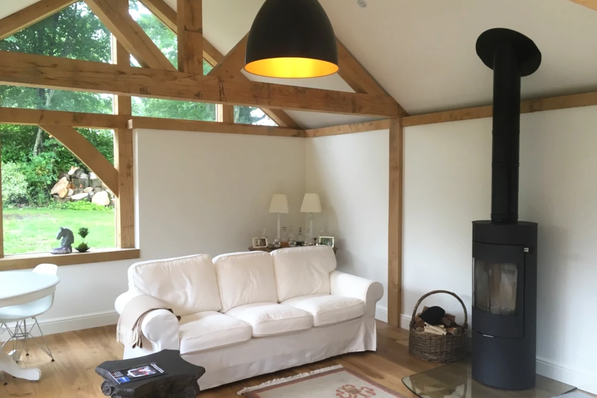 Modern oak frame living room featuring exposed beams, wood burning stove, white sofa and vaulted ceiling with garden views through feature windows