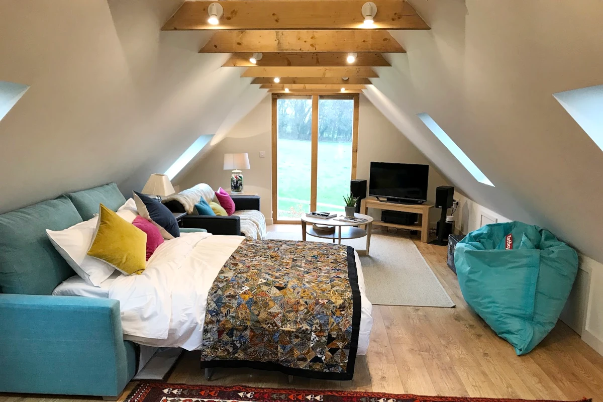 Modern loft conversion featuring exposed oak beams, full-height window, comfortable seating area and entertainment space with natural light from velux windows