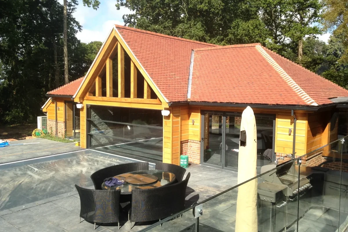 Oak framed pool house featuring full-height glass panels, clay-tiled roof, and slate terrace with rattan furniture set against a leafy backdrop