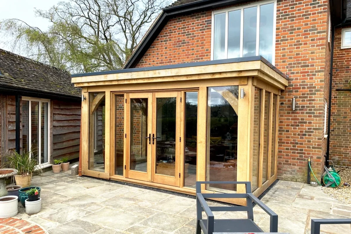 Handcrafted oak framed orangery featuring bi-fold doors, glazed panels and flat roof lantern, complementing red brick house with stone patio