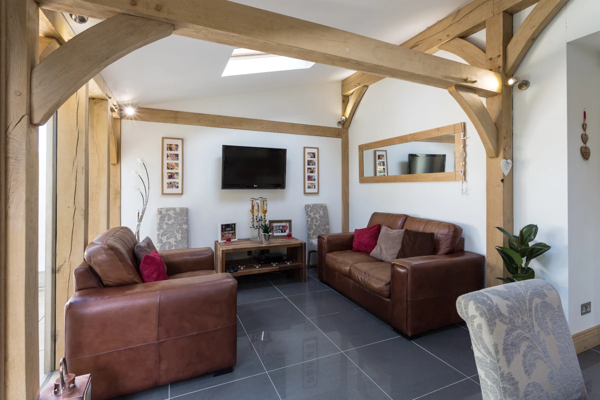 Oak-framed living room featuring exposed beams, leather sofas, grey floor tiles and wall-mounted tv with natural light from skylights