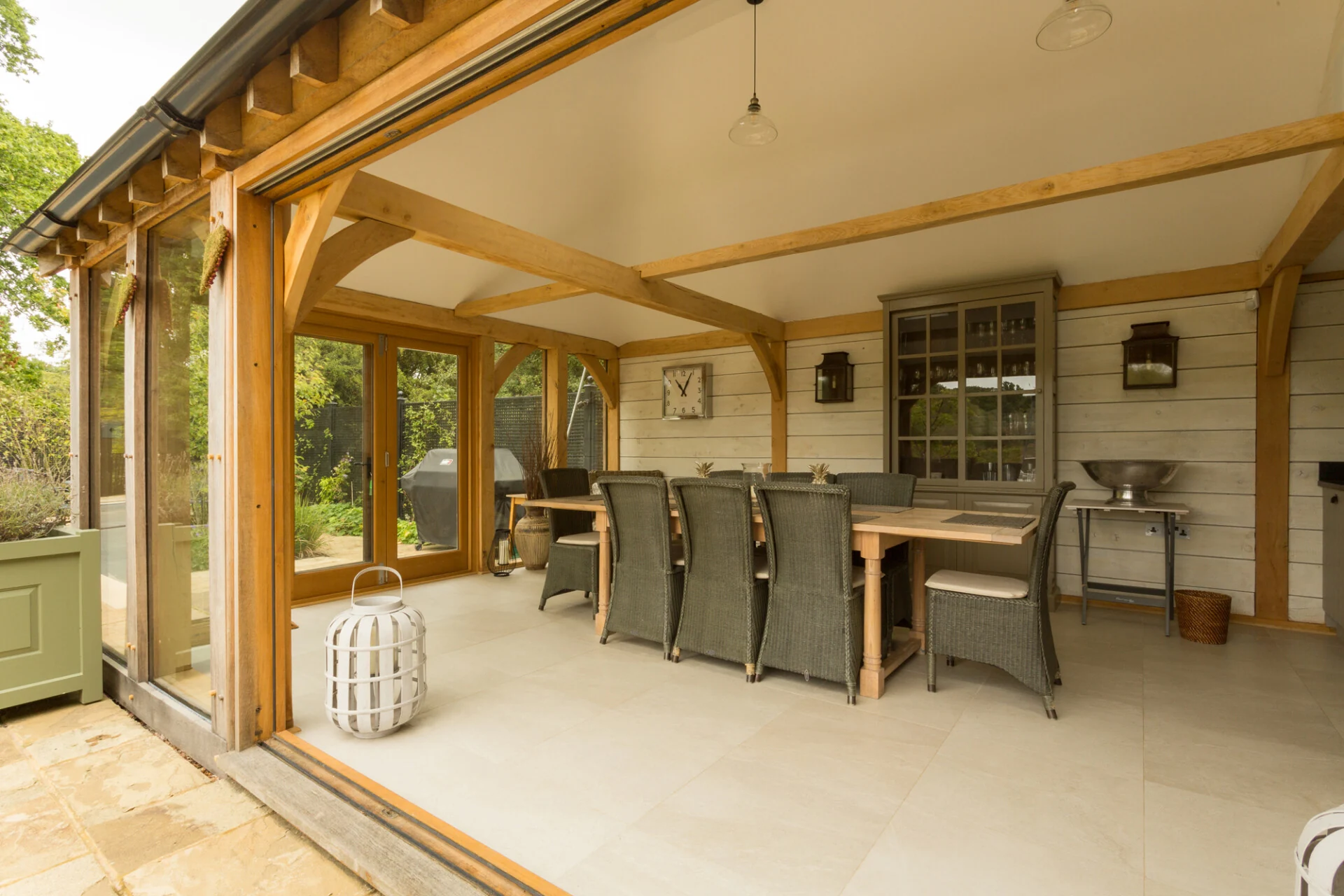 Oak frame garden room featuring curved braces, limestone flooring, weatherboard cladding and glazed panels, furnished with oak dining table and lloyd loom chairs