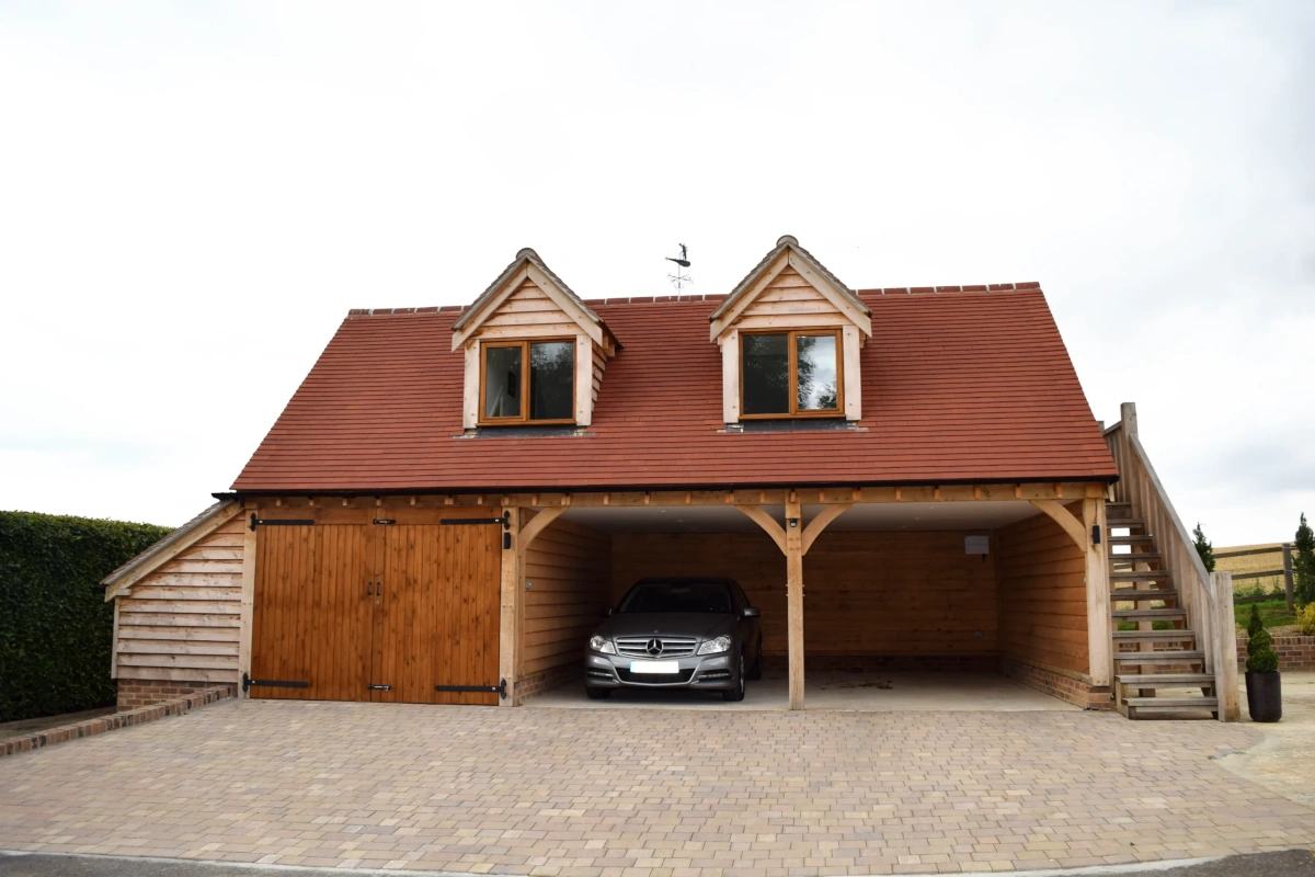 Oak-framed three-bay garage featuring twin dormer windows, enclosed storage bay, and open car port with mercedes parked beneath