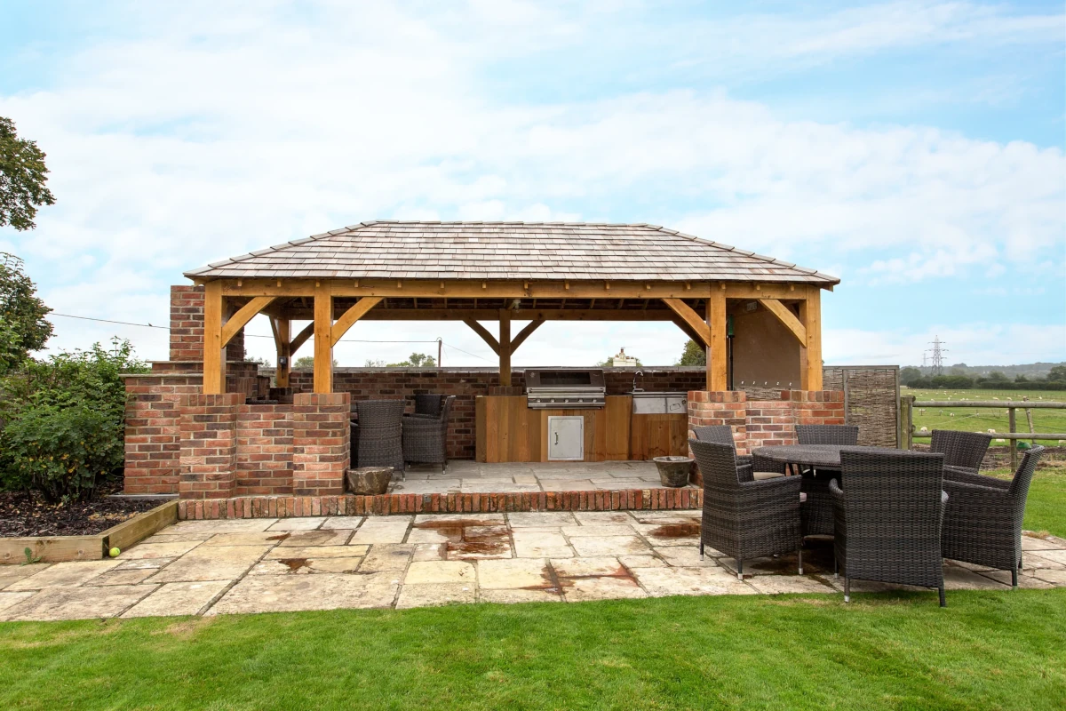 Oak gazebo with outdoor kitchen featuring brick walls, cedar-shingled roof, built-in barbecue and rattan dining furniture overlooking pastoral landscape