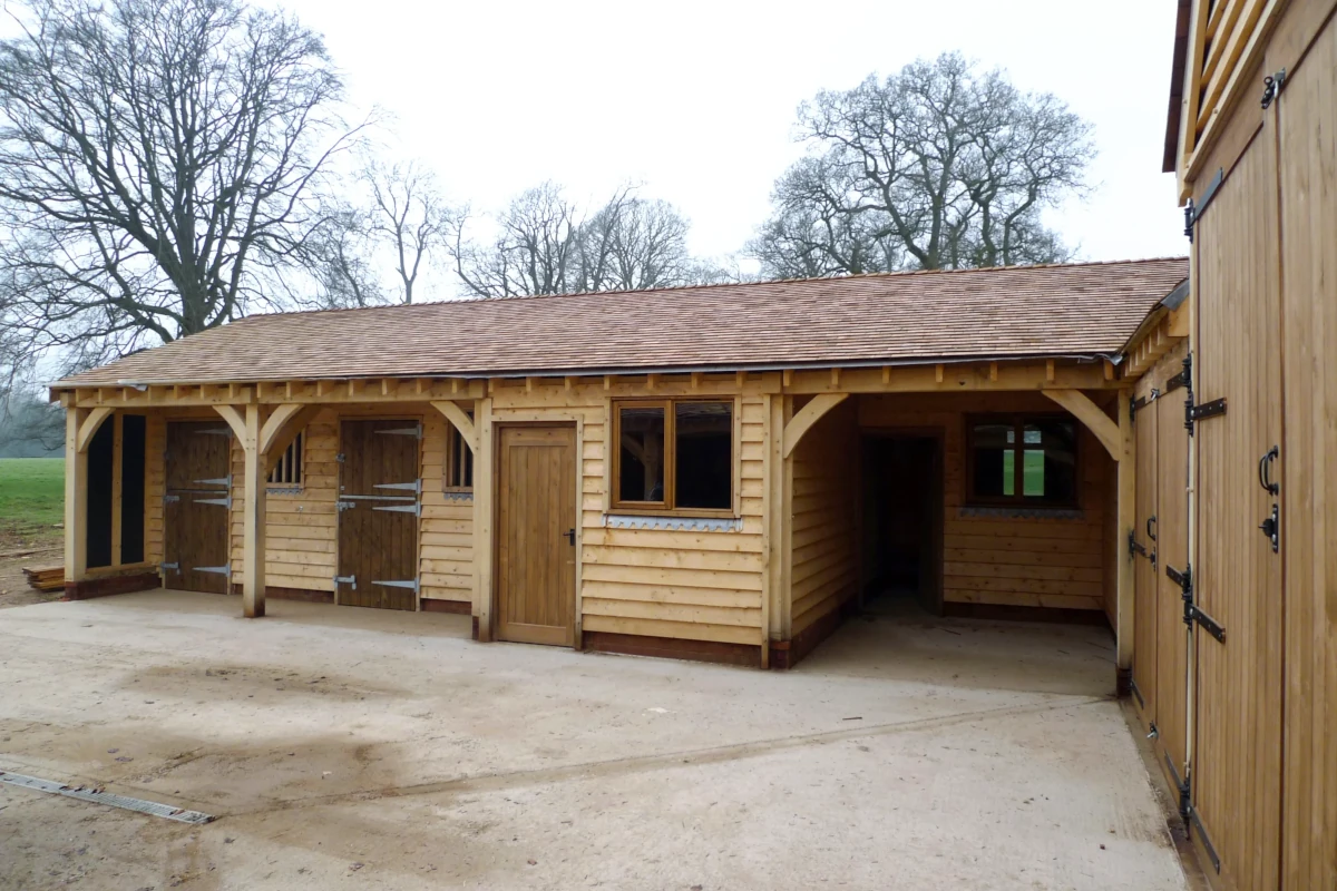 Oak framed stable block featuring two horse boxes, central tack room with window, weatherboarding and clay-tiled roof set against winter trees