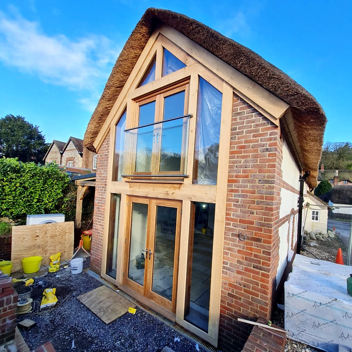 Oak framed extension featuring double-height glazing, thatched gable roof and traditional brick walls under construction, oak framing from oakcraft's hampshire workshop