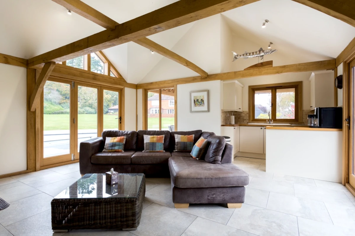 Interior of luxury oak framed pool house featuring vaulted ceiling with exposed beams, full-height glazing, modern kitchen and comfortable living area