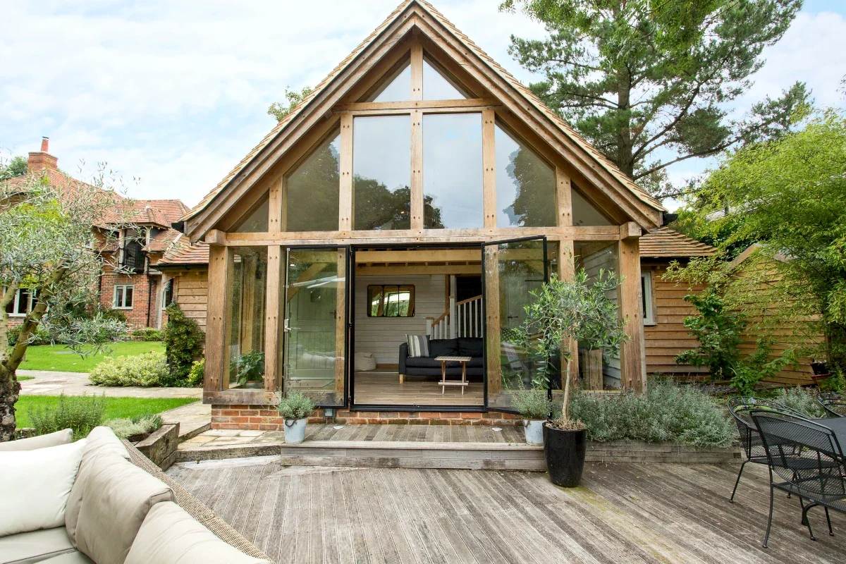 Bespoke Oak Framed Buildings In Hampshire Bespoke oak framed annexe in Hampshire featuring vaulted ceiling with full-height glazed gable end, timber cladding, bi-fold doors opening onto wooden decking with Mediterranean-style planting, handcrafted by OakCraft