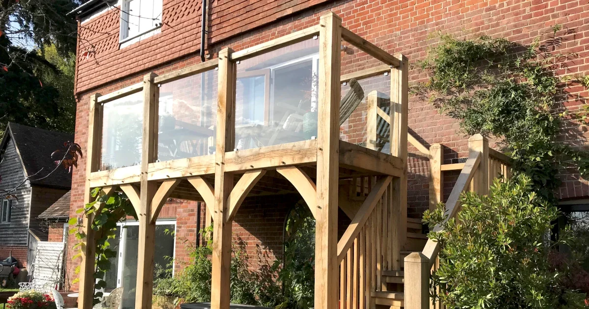 Oak Framed Balconies Bespoke oak framed balcony with glass panels and exterior wooden staircase featuring traditional curved braces on a Sussex red brick home surrounded by mature garden