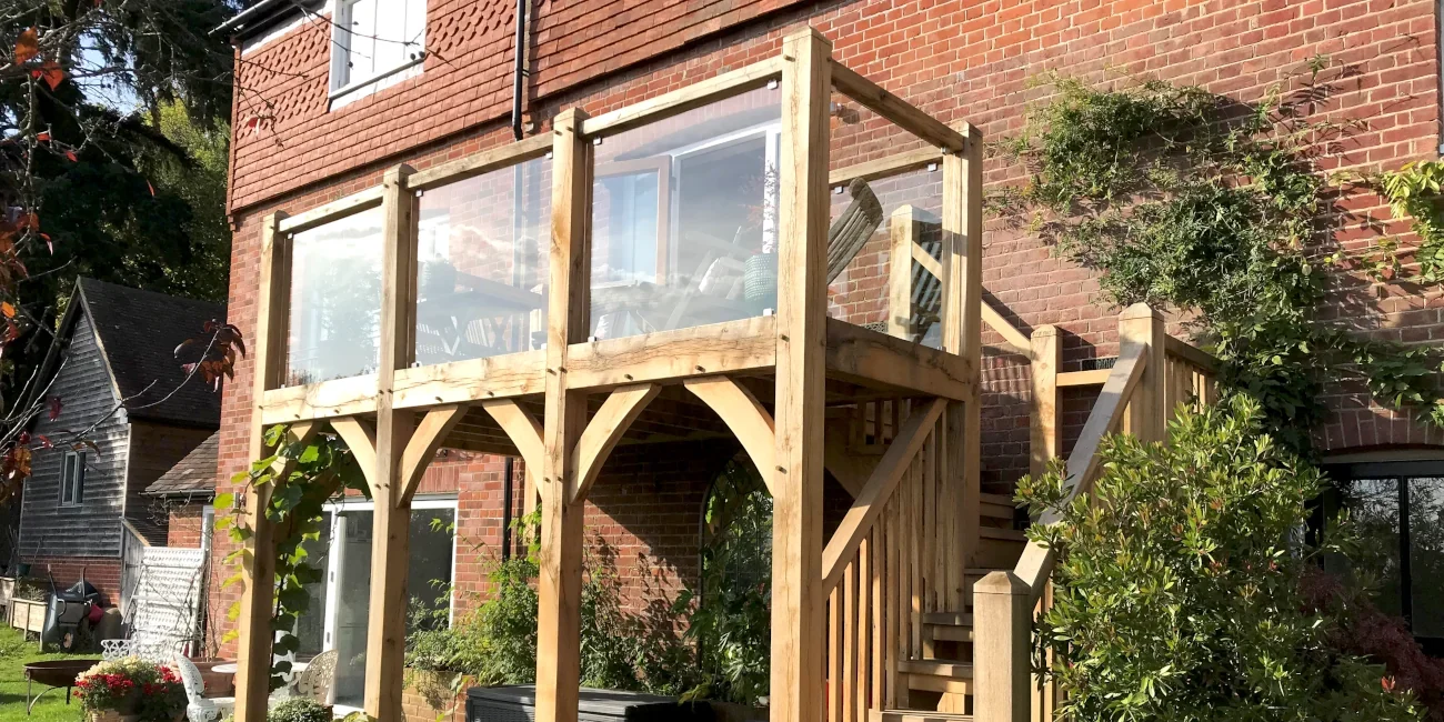 Oak Framed Balconies Handcrafted oak framed balcony with glass panels and timber staircase featuring traditional curved braces attached to red brick family home in Surrey
