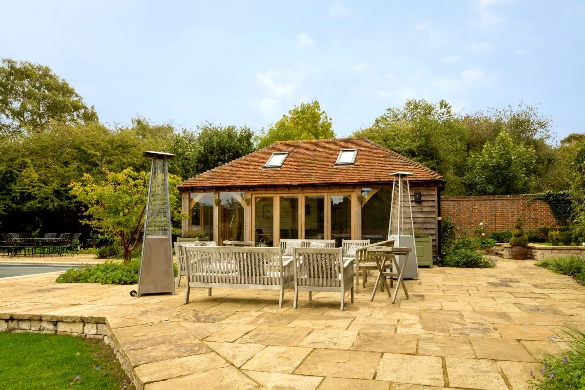Bespoke Oak Framed Buildings In Hampshire Handcrafted bespoke oak framed pool house and garden room in Hampshire with clay tiled roof, floor-to-ceiling glazing and Yorkstone terrace featuring outdoor furniture and patio heaters