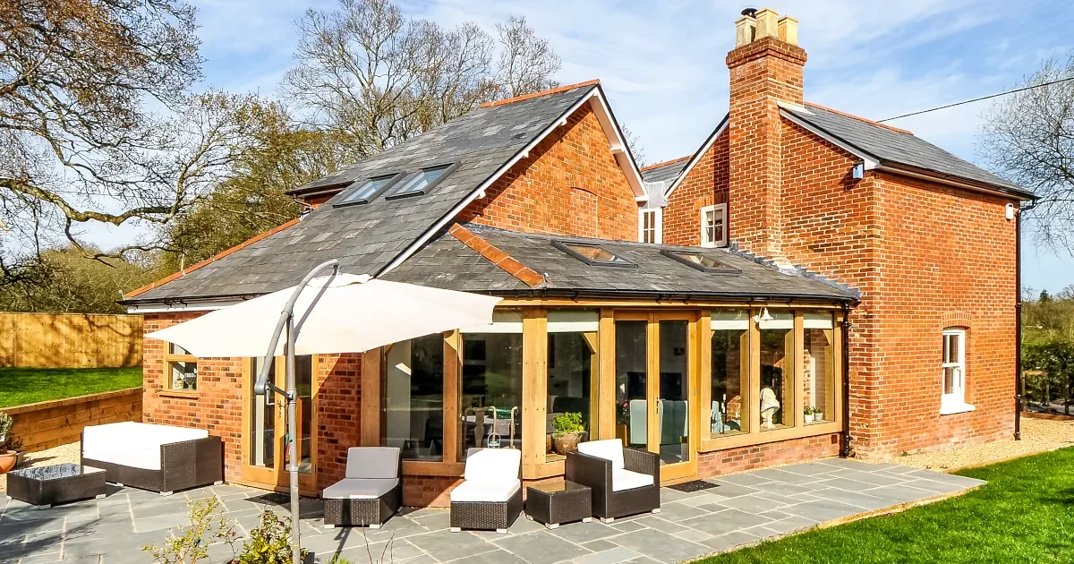 Oak Framed Buildings Hampshire Stunning oak framed buildings Hampshire showcase: traditional red brick cottage with bespoke oak frame extension featuring natural slate roof, skylights, full-width glass doors opening onto stone patio with rattan furniture, set in picturesque countryside among mature trees