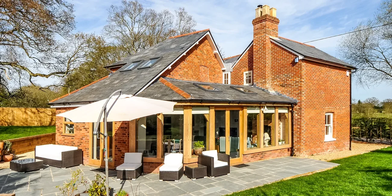 Oak Framed Buildings Hampshire Red brick cottage in Hampshire countryside featuring bespoke oak framed extension with natural slate roof, skylights, full-width glass doors, grey stone terrace with rattan garden furniture and cantilever parasol, surrounded by mature trees and landscaped garden