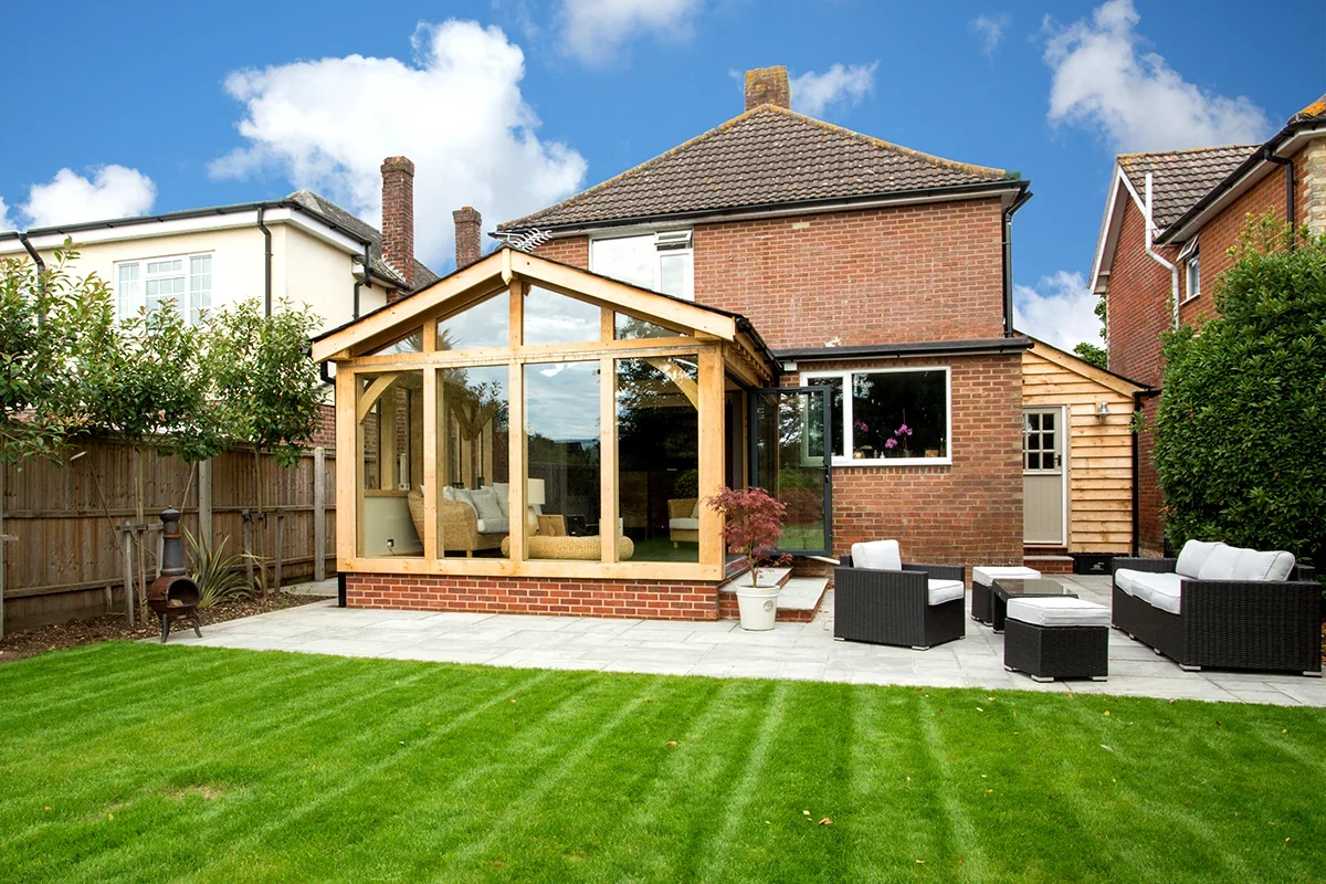 Oak Framed Buildings Hampshire Stunning oak framed garden room extension with full-height glazing on a traditional Hampshire brick house, featuring a manicured lawn and stone patio with rattan garden furniture