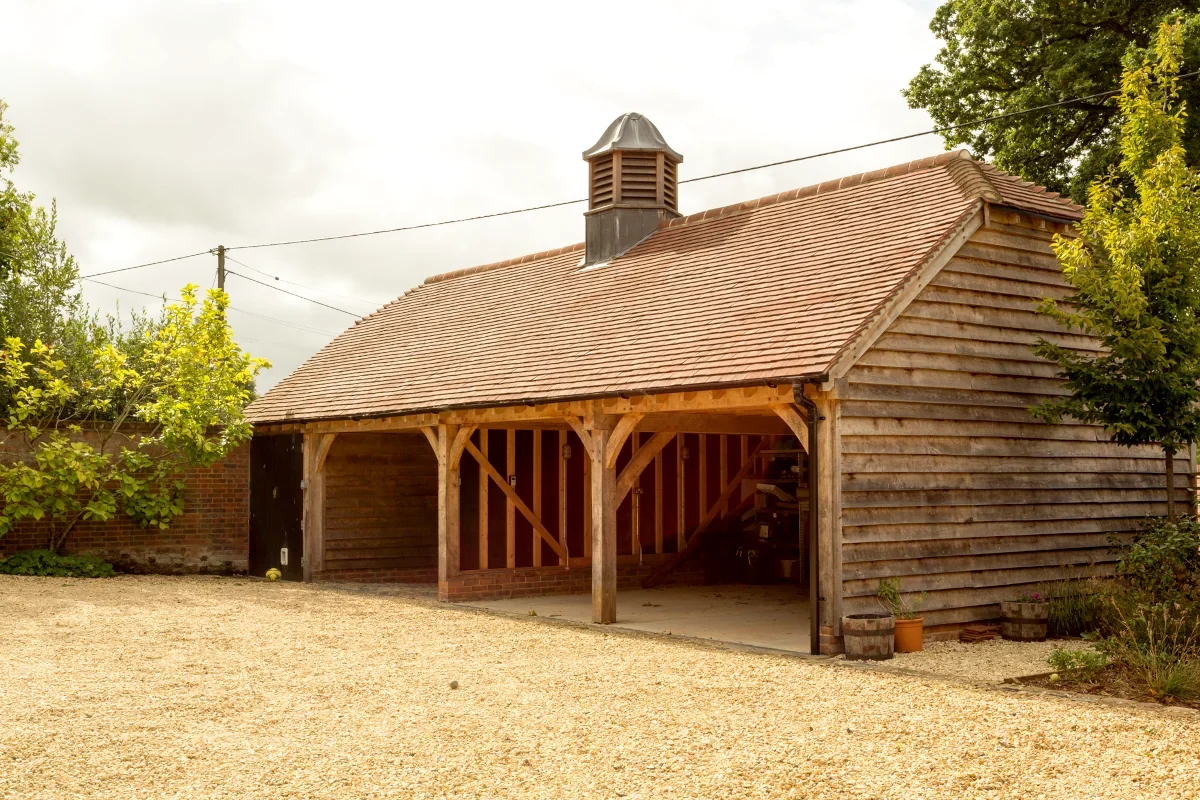 Oak Framed Buildings Hampshire Handcrafted oak framed garage in Hampshire featuring weathered timber cladding, clay tile roof, oak posts, cupola ventilation, gravel driveway and adjoining brick wall